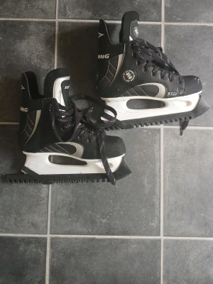 Svarta skridskor med vita detaljer - Säljer ett par svarta skridskor med vita detaljer och snörning. Skridskorna har robusta bladskydd och hög ankel för extra stöd. Perfekta för isträning eller hockey. Snygg sportig design som passar både nybörjare och mer erfarna åkare.