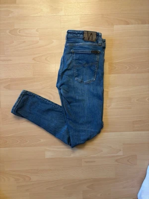 Nudie Jeans - Blå  - Snygga jeans från Nudie Jeans Co i en klassisk blå tvätt. Stilren modell med snygga detaljer och Nudie-loggan baktill. Jeansen är i fint begagnat skick med naturliga slitningar som ger dem en snygg och avslappnad look. 📏 Storlek: W32 L30 👖 Färg: Blå denim 📦 Skickas snabbt och välpackat!