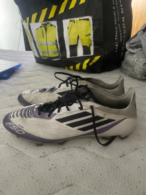 Adidas F50 Messi fotbollsskor lila/vit - Säljer ett par Adidas F50 Messi fotbollsskor i vitt och lila med svarta detaljer. Skorna har snörning och låg profil, samt klassiska tre ränder på sidan. Designade för fart på planen och tillverkade i syntetmaterial för bra bollkontroll.