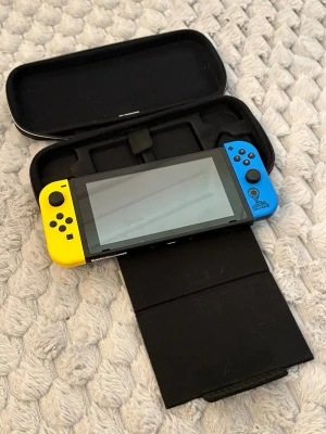 Nintendo Switch med fodral - Nintendo Switch spelkonsol med en gul Joy-Con till vänster och en blå Joy-Con till höger. Konsolen har en stor skärm och medföljer ett svart skyddsfodral i hårdplast och tyg. Perfekt för gaming både hemma och på språng. Det följer även med två spel, NBA2k26 och Pokémon sword.