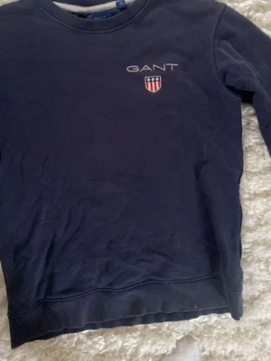 Mörkblå sweatshirt från GANT - Mörkblå sweatshirt från GANT med rund halsringning och broderad logga samt sköld på bröstet. Klassisk modell med ribbade muddar vid ärmslut och nederkant. Perfekt till jeans eller chinos för en clean look. Skulle säga att modellen passar bättre till pojkar 12-14 i och med att den är lite liten.