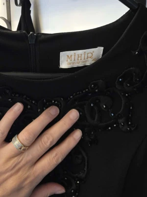 Svart långklänning med pärldetaljer - Elegant svart långklänning från Mihir med vackra pärldetaljer och broderier upptill. Klänningen har en rund halsringning och dragkedja bak. Tyget är lätt och faller snyggt, perfekt för speciella tillfällen.