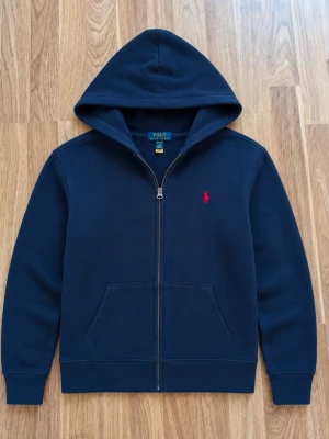 Ralph Lauren zip hoodie - Säljer en marinblå hoodie från Polo Ralph Lauren med röd broderad logga på bröstet. Klassisk modell med dragkedja, huva och fickor framtill. Perfekt till både vardag och en mer stilren look. • Färg: Marinblå • Märke: Polo Ralph Lauren • Modell: Zip-up hoodie • Skick: Bra skick, normal användning • Mjuk och bekväm passform