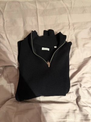 Svart stickad tröja från Jack & Jones - Snygg svart stickad tröja från Jack & Jones med halv dragkedja och hög krage. Tröjan är ribbstickad och har en stilren look som passar perfekt till jeans eller chinos. Materialet känns mjukt och bekvämt mot huden.