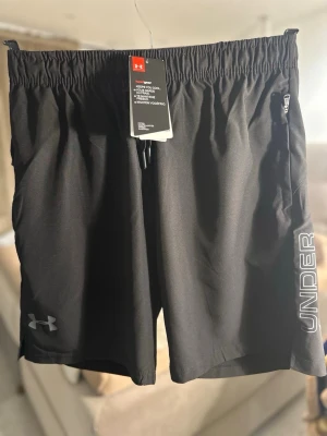 Svarta träningsshorts från Under Armour - Snygga svarta träningsshorts från Under Armour med elastisk midja och dragsko. Shortsen har logga på vänster ben och texten 'UNDER ARMOUR' längs sidan. Praktisk ficka med dragkedja på höger sida. Perfekta för gymmet eller löprundan. Finns även L