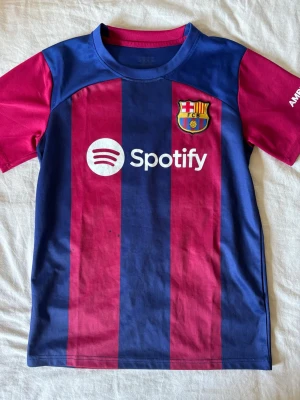 Barcelona matchtröja Lamine Yamal 27 - Säljer en officiell FC Barcelona matchtröja med Lamine Yamal och nummer 27 på ryggen. Tröjan har breda blå och röda ränder, korta ärmar och tryckta loggor från Spotify och UNHCR. Perfekt för fotbollsfans som vill stötta sitt lag.