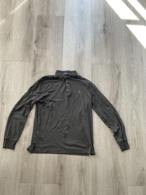 Grå pikétröja från Polo Ralph Lauren - Säljer en grå pikétröja från Polo Ralph Lauren med klassisk krage och knappar framtill. Tröjan har den ikoniska broderade polospelaren på bröstet och är i en slim fit-modell. Perfekt för en clean och stilren look.