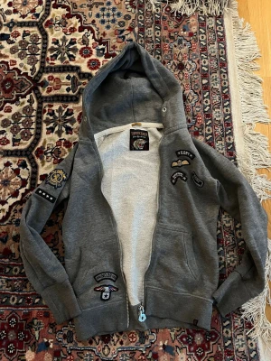 Grå SUPERDRY hoodie - Grå hoodie med dragkedja och flera coola patchar på bröstet och ena ärmen. Hoodien har två fickor framtill och ribbade muddar vid ärmslut och nederkant. Insidan är mjuk och skön, perfekt för chill dagar. Märket 'Superior' syns i nacken.