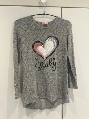 Grå långärmad topp med paljett-hjärta - Grå långärmad topp med stort hjärta i paljetter och fluffigt tyg framtill. Texten 'Baby' och små hjärtan tryckta under motivet. Mjuk och bekväm, perfekt för dig som gillar gulliga detaljer.
