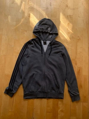 Mörkgrå zip hoodie från Adidas - Mörkgrå hoodie från Adidas med dragkedja, huva med svarta snören och klassiska svarta ränder längs ärmarna. Broderad Adidas-logga på bröstet och två stora fickor framtill. Tillverkad i mjukt sweatshirtmaterial, perfekt för chill dagar.