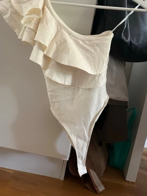 Krämvit body med volanger - Snygg krämvit body med asymmetrisk axel och dubbla volanglager framtill. Ena axeln är bar och den andra har en smal axelrem. Tillverkad i mjukt, stretchigt tyg och knäpps med tryckknappar i grenen.