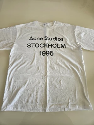 Vit Acne Studios t-shirt - Vit t-shirt från Acne Studios med svart texttryck 'STOCKHOLM 1996' på bröstet. Klassisk passform och rund halsringning. Perfekt basplagg för en clean och stilren look.