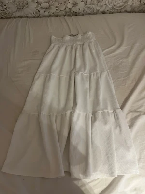 Vit långkjol med volanger - Säljer en vit långkjol med bred resår i midjan och flera volanglager som ger ett flowy och luftigt intryck. Kjolen har en enkel och stilren look, perfekt för dig som gillar en avslappnad men ändå trendig stil.