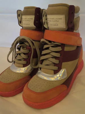 Marc Jacobs färgglada high top sneakers - Unika high top sneakers från Marc Jacobs i beige, orange och vinrött med holografiska detaljer. Skorna har snörning, kardborreband och en röd sula. Tillverkade i en mix av mocka och läder för en cool och trendig look.