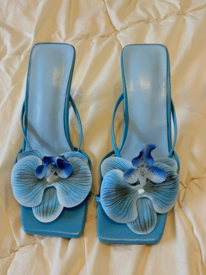 Blå pumps med orkidédekor - Unika blå pumps med fyrkantig tå och smal klack. Skorna har en stor 3D-blomma i form av en blå orkidé framtill som ger en riktigt statement-look. Tillverkade i glansigt material med öppen häl och tunna remmar.