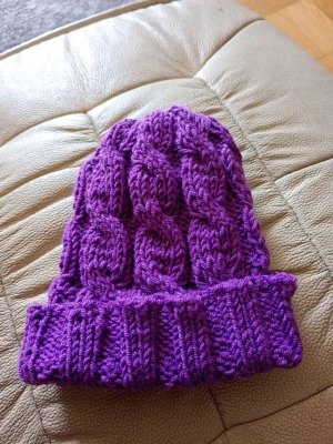 Lila kabelstickad mössa - Säljer en lila kabelstickad mössa med uppvikt kant. Mössan är handstickad i tjockt garn och har ett snyggt flätmönster som ger en cool och mysig look. Perfekt för kalla dagar när du vill hålla dig varm och ändå se trendig ut.