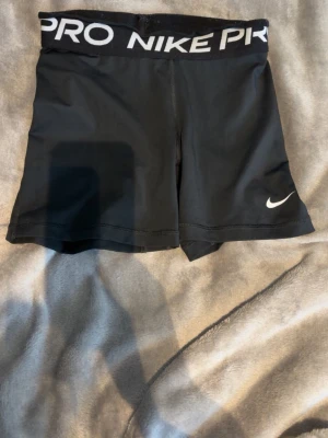 Svarta Nike Pro träningsshorts - Lite vikt längst upp i kanten, går säkert att fixa. Säljer ett par svarta Nike Pro shorts med bred resår i midjan där det står 'NIKE PRO' i vitt. Shortsen har en tight passform och är perfekta för träning. Materialet är stretchigt och känns bekvämt mot huden. Klassisk Nike-logga på benet.