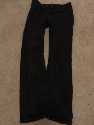 Svarta bootcut jeans - Säljer ett par svarta bootcut jeans med klassisk femficksdesign och knappgylf. Jeansen har raka ben och subtila slitningar för en avslappnad look. Perfekta till sneakers eller boots och passar dig som gillar en tidlös stil.