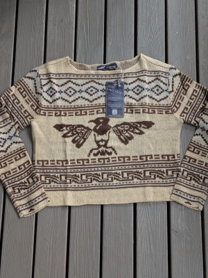 Beige och brun stickad tröja Arizona  - Stickad tröja i beige med brunt mönster från Arizona. Tröjan har ett stort örnmotiv framtill och geometriska mönster på ärmar och kropp. Rund halsringning och långa ärmar. Perfekt för dig som gillar unika stickade plagg med retrovibbar.
