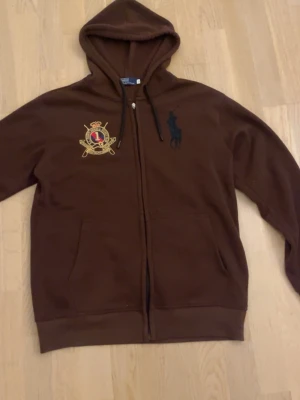 Brun hoodie från Ralph Lauren - Brun hoodie med dragkedja från Ralph Lauren. Stora broderade loggor på bröstet och ryggen, samt huva med dragsko. Mjuk bomullskänsla och två fickor framtill. Perfekt för en avslappnad och stilren look.