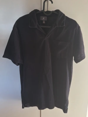 Svart kortärmad skjorta från H&M - Svart skjorta från H&M i regular fit med korta ärmar och klassisk krage. Skjortan har en bröstficka och är tillverkad i ett mjukt material som känns bekvämt mot huden. Perfekt för en avslappnad och stilren look.