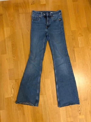 Blå bootcut jeans med hög midja - Ett par klassiska blå bootcut jeans med hög midja och fem fickor. Jeansen har utsvängda ben och är gjorda i ett mjukt denimtyg. Perfekta för dig som gillar retrostil och vill ha ett par bekväma jeans med snygg passform.