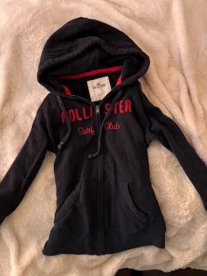 Svart Hollister hoodie med dragkedja - Mörkblå vintage hollister zip tröja/kofta med rött tryck o fint skick! Stl L men liten i storlek mer som en S❣️