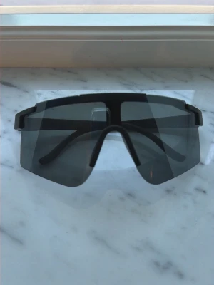 Svarta sportiga solglasögon - Säljer ett par svarta solglasögon med sportig och futuristisk design. De har stora, rektangulära linser som täcker mycket av ansiktet och en bred båge i svart. Perfekta för dig som vill ha en cool och modern look till sommaren.