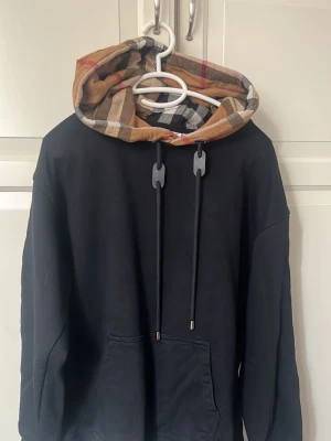 Svart hoodie med rutmönstrad huva - Snygg svart hoodie med stor ficka framtill och en huva i klassiskt beige, rött och svart rutmönster. Hoodien har långa ärmar och svarta dragsnören med metalländar. Perfekt för en avslappnad streetwear-look.