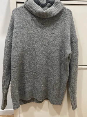 Grå polotröja i stickat material - Mysig grå stickad polotröja med avslappnad passform och långa ärmar. Tröjan har en bred, vikt polokrage och är tillverkad i ett mjukt, värmande material som passar perfekt för kyliga dagar. Enkel och stilren design som är lätt att matcha.