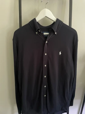 Svart skjorta från Polo Ralph Lauren - Klassisk svart skjorta från Polo Ralph Lauren med button-down krage och vit broderad logga på bröstet. Tillverkad i lätt bomullsmaterial och har knappar framtill. Perfekt för en clean och stilren look.