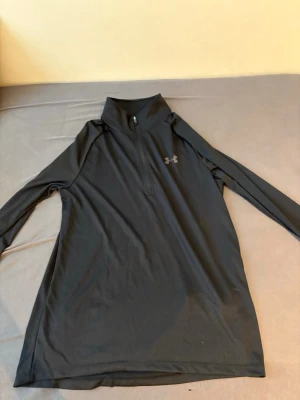 Svart träningströja Under Armour - Svart långärmad träningströja från Under Armour med halv dragkedja framtill och hög krage. Tillverkad i ett stretchigt funktionsmaterial som andas, perfekt för löpning eller gym. Diskret logga på bröstet och ryggen.