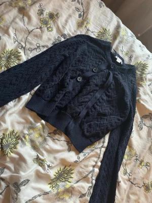 Mörkblå stickad kofta Gina Tricot - Snygg mörkblå stickad kofta från Gina Tricot med dekorativa knappar framtill och ribbade muddar. Koftan har ett fint hålmönster och är gjord i mjukt material. Perfekt att slänga över en topp för en trendig look.