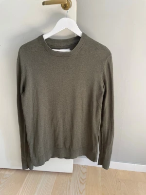 Olivgrön/grön volt tröja som passar bra till blåa jeans - En enkel och stilren olivgrön tröja med rund halsringning och långa ärmar. Tröjan är tillverkad i mjuk bomull och har ribbade muddar vid hals, ärmslut och nederkant. Perfekt för lager på lager och passar till det mesta.