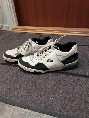 Vita  Lacoste sneakers - Säljer ett par vita och svarta sneakers från Lacoste med klassisk krokodil-logga på sidan. Skorna har lågt skaft, snörning och detaljer i svart runt sulan och hälen. Materialet är en mix av läder och syntet. Perfekta för dig som gillar sportig och clean stil. Skriv vid frågor. Äkta såklart.