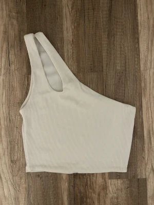 Vit oneshoulder ribbad topp från Pull & Bear - Vit oneshoulder topp i ribbat material i storlek S från Pull & Bear. Toppen är ärmlös och har en asymmetrisk axelbandslös design. 