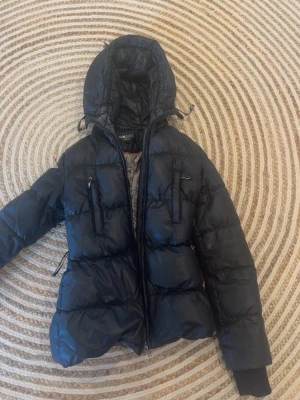 Svart pufferjacka med huva - Säljer en svart pufferjacka med stor huva och dragkedja framtill. Jackan har två bröstfickor med dragkedja och justerbara detaljer vid huvan. Perfekt för kalla vinterdagar och riktigt skön att bära tack vare det vadderade materialet.