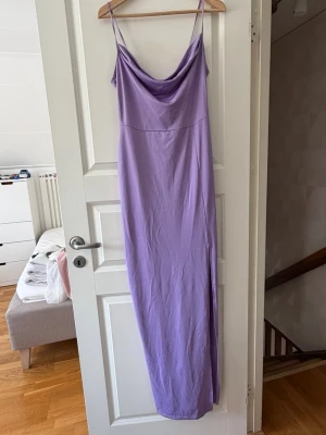 Lång lila klänning med slits - Säljer en lång, lila klänning med tunna axelband och snygg draperad urringning. Klänningen har en slits på sidan som ger extra rörelsefrihet och en elegant look. Perfekt för sommarens alla tillfällen!