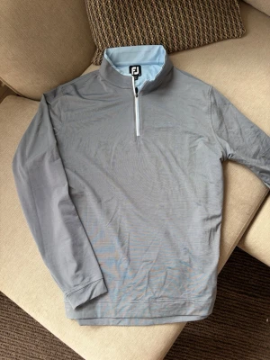 FootJoy 1/4 Zip Golftröja – grå/blå, storlek L - Säljer en stilren långärmad golftröja från FootJoy i grått med ljusblå detaljer. Modellen har kort dragkedja framtill, hög krage och ett diskret sportigt uttryck som passar perfekt både på golfbanan och till vardags.  Storlek L. Tröjan är iprincip ny och har snygga detaljer som kontrastfärgad insida i kragen, ljusblå kantning samt broderad FJ-logga baktill.  Bekväm och lätt modell som fungerar bra som mellanlager under svalare rundor. 
