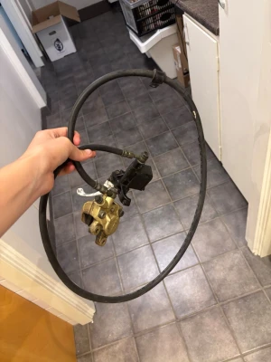 Bromsok med slang till cykel eller moped - Säljer ett bromsok i metall med tillhörande bromsslang, troligen för cykel eller moped. Metallfärgad med svart slang och fäste. Perfekt för dig som behöver reservdelar till ditt fordon.