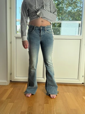 Blå bootcut jeans med råa kanter - Säljer ett par blå bootcut jeans med markerad tvätt och råa, fransiga kanter längs sidorna och vid bensluten. Jeansen har hög midja, klassiska fem fickor och stängs med dragkedja och knapp. Perfekta för en trendig och avslappnad look.
