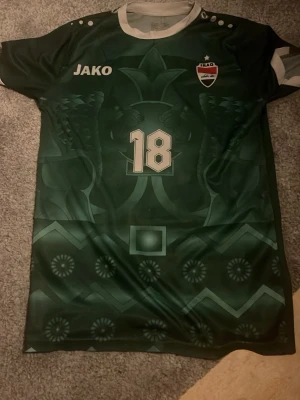 Grön Irak fotbollströja Jako #18  - Priset kan diskuteras. Säljer en grön fotbollströja från Jako med Iraks landslagsemblem och nummer 18 på både fram- och baksidan. Tröjan har vita detaljer, rund hals och trycket 'AYMEN' på ryggen. Tillverkad i lätt och ventilerande material, perfekt för träning eller match.