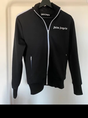 Svart hoodie från Palm Angels - Snygg svart hoodie från Palm Angels med vit dragkedja och logga på bröstet. Hoodien har långa ärmar, två fickor med dragkedja och en klassisk passform. Materialet är mjukt och bekvämt, perfekt för en avslappnad streetwear-look.