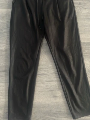 Svarta leggings i fuskskinn - Säljer ett par svarta leggings i fuskskinn med hög midja och resår i midjan. De har en snygg glansig finish och smal passform som ger en trendig look. Perfekta att styla med oversized tröja eller crop top.