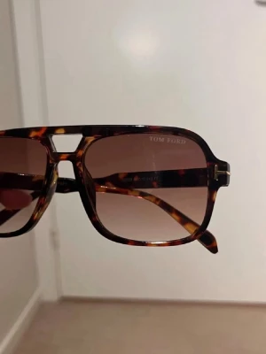Tom Ford bruna solglasögon - Snygga solglasögon från Tom Ford med brunmelerade bågar i klassisk aviatorform. Glasen är bruna och bågarna har en sköldpaddsmönstrad look som ger en exklusiv känsla. Perfekta för dig som vill ha en trendig accessoar med lyxig touch.