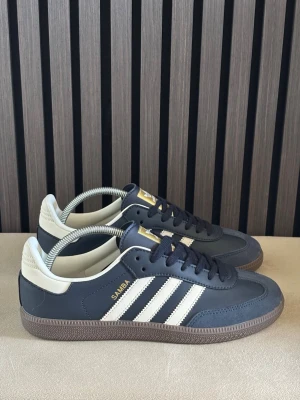 Adidas Samba mörkblå sneakers - Snygga Adidas Samba sneakers mörkblått och vitt med klassiska tre ränder på sidan och gulddetaljer. Ovandelen är i läder och mocka, med vit häl och brun gummisula. Perfekta för dig som gillar retrostil och streetwear.