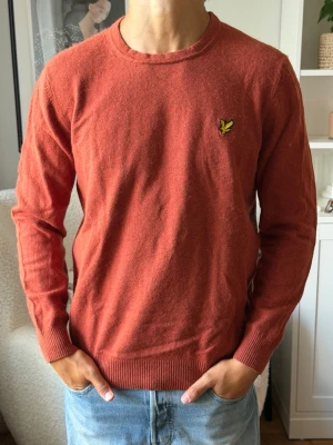 Rostfärgad stickad tröja Lyle & Scott - Snygg rostfärgad stickad tröja från Lyle & Scott med rund halsringning och gul logga på bröstet. Tröjan har långa ärmar och ribbade muddar vid ärmslut och nederkant. Perfekt till jeans för en avslappnad look. Sitter som en M