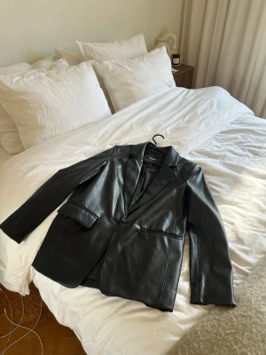 Svart kavaj i skinnimmitation  - Snygg svart skinnjacka med klassisk krage och knappstängning. Perfekt för dig som vill ha en stilren och trendig look. Passar till många olika outfits.