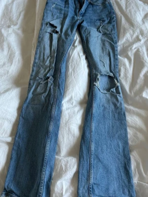 Blå slitna bootcut jeans - Säljer ett par ljusblå bootcut jeans med flera slitningar och hål på benen. 