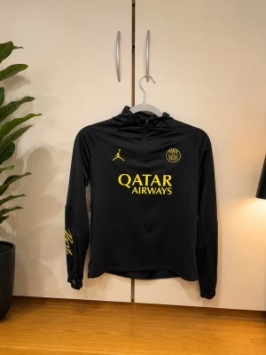 Svart PSG träningströja  - Snygg svart träningströja från PSG x Jordan med huva och halv dragkedja. Tröjan har gula detaljer, PSG-logga och Qatar Airways-tryck på bröstet samt Jordan-logga. Långa ärmar med meshpaneler och tryck på ärmen.
